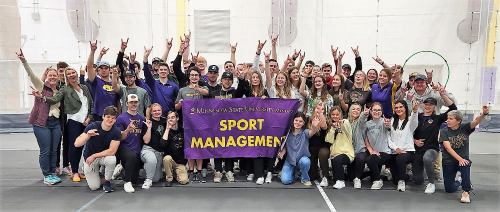 A large group of students from HAN University and Minnesota State University posing while doing the horns up with their hands and holding a Minnesota State University, Mankato Sport Management banner in Myers Field House
