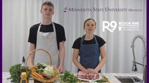 Two Region Nine dietetics students standing in front of a white screen and behind a cooking table prepping food to film a video
