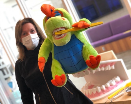 Dental hygiene faculty holding an educational dinosaur puppet brushing its teeth in the dental clinic lobby with the giant mounth model on a table in the background