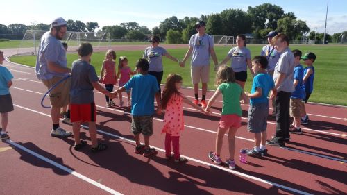 Health and physical education students in a circle holding hands with children outside on the track during a kids activity
