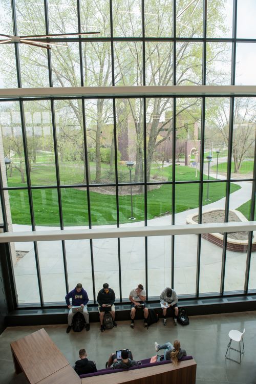 Interior photo of large wall of windows with several students studying with laptops in the main lobby of the College of Allied Health and Nursing building