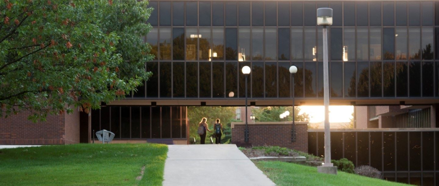 Two students walking between the Trafton buildings with the sun shining towards the students as it is setting