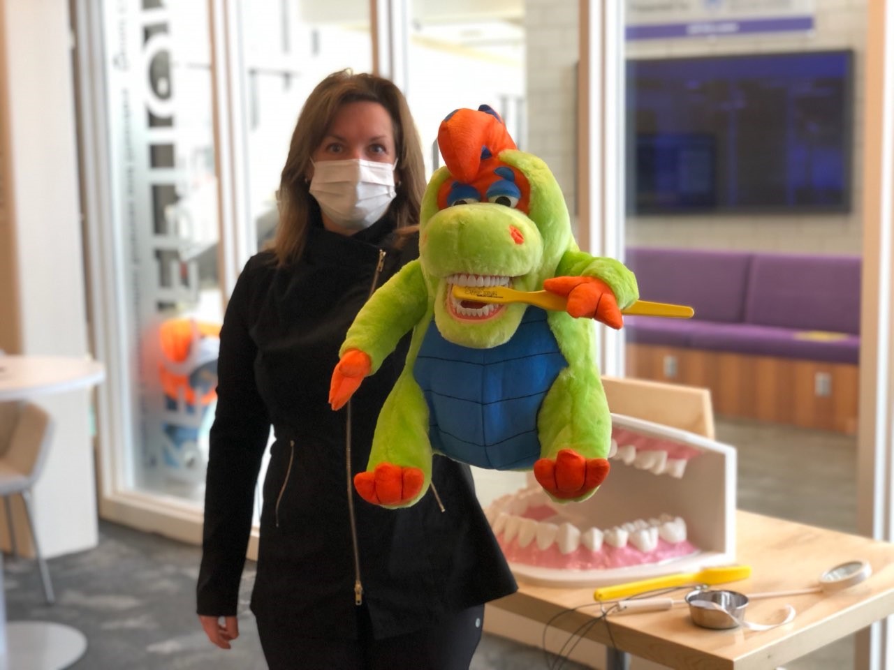 Faculty in dental clinic lobby with green dinosaur puppet designed to help kids learn to brush their teeth