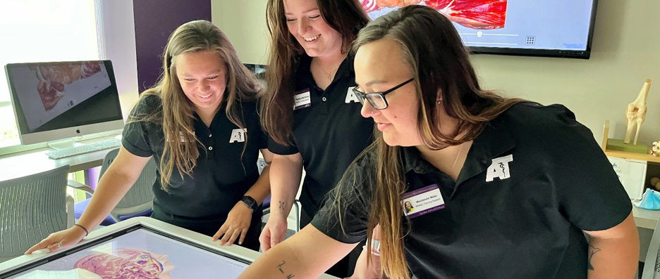 Three athletic training students studying the muscle structure of the human body at a table screen in the athletic training lab