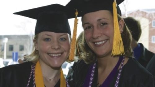 Two students in graduation cap and gown posing in the hallway with smiles