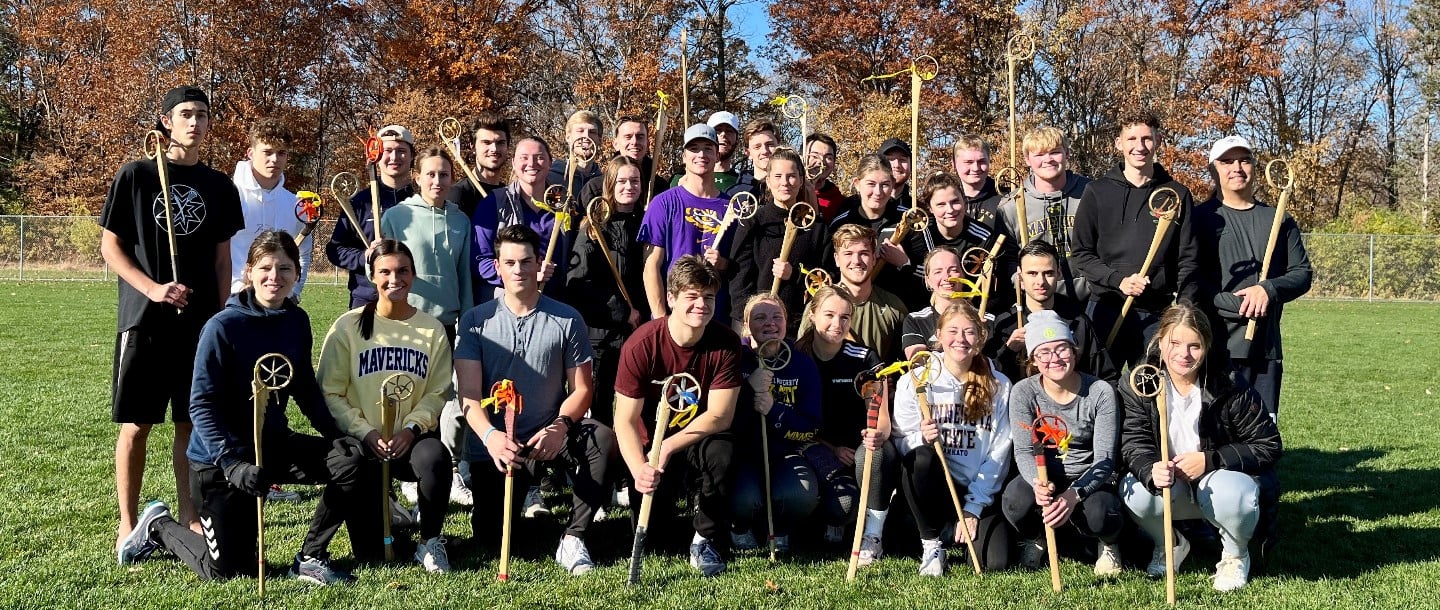 A large group of Sport Management students outside on a lacrosse field with many holding wooden lacrosse sticks of different colors