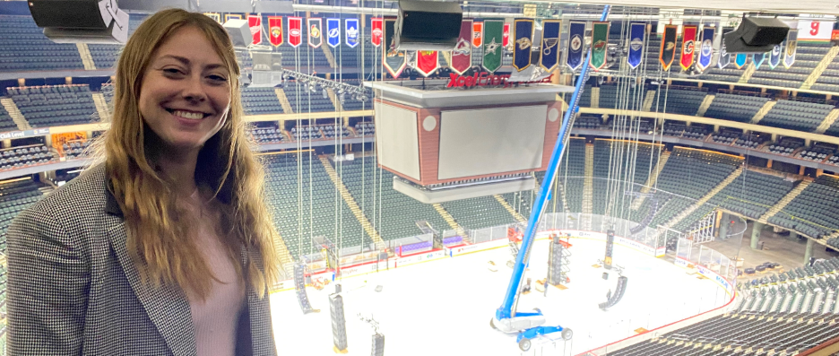 Sport Management student posing inside of the Excel Energy Center during the set up for a Minnesota Wild hockey game with the arena and score board in the background