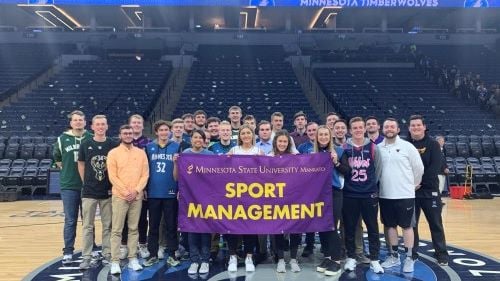 A group of Sport Management students posing on the Minnesota Timerwolves basketball court holding the Sport Management banner