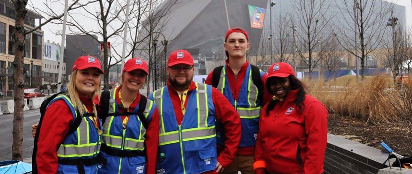 Five students of Bachelor's of Sports Management outside of a stadium in red shirt and cap and blue and neon colored safety vests