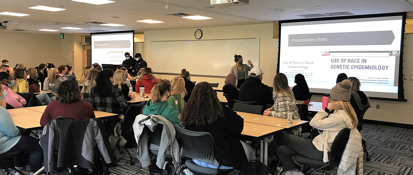 Student wearing masks at IDEAS workshop about the "Use of Race in Genetic Epidemiology" with faculty presenting and two more students in front of the computer