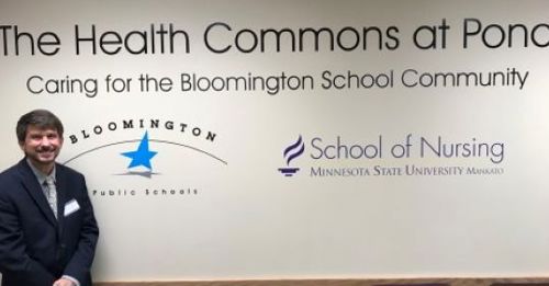 Wm. Eric Strong in front of The Health Commons at Pond sign that includes the Bloomington Public Schools and Minnesota State Mankato School of Nursing logos