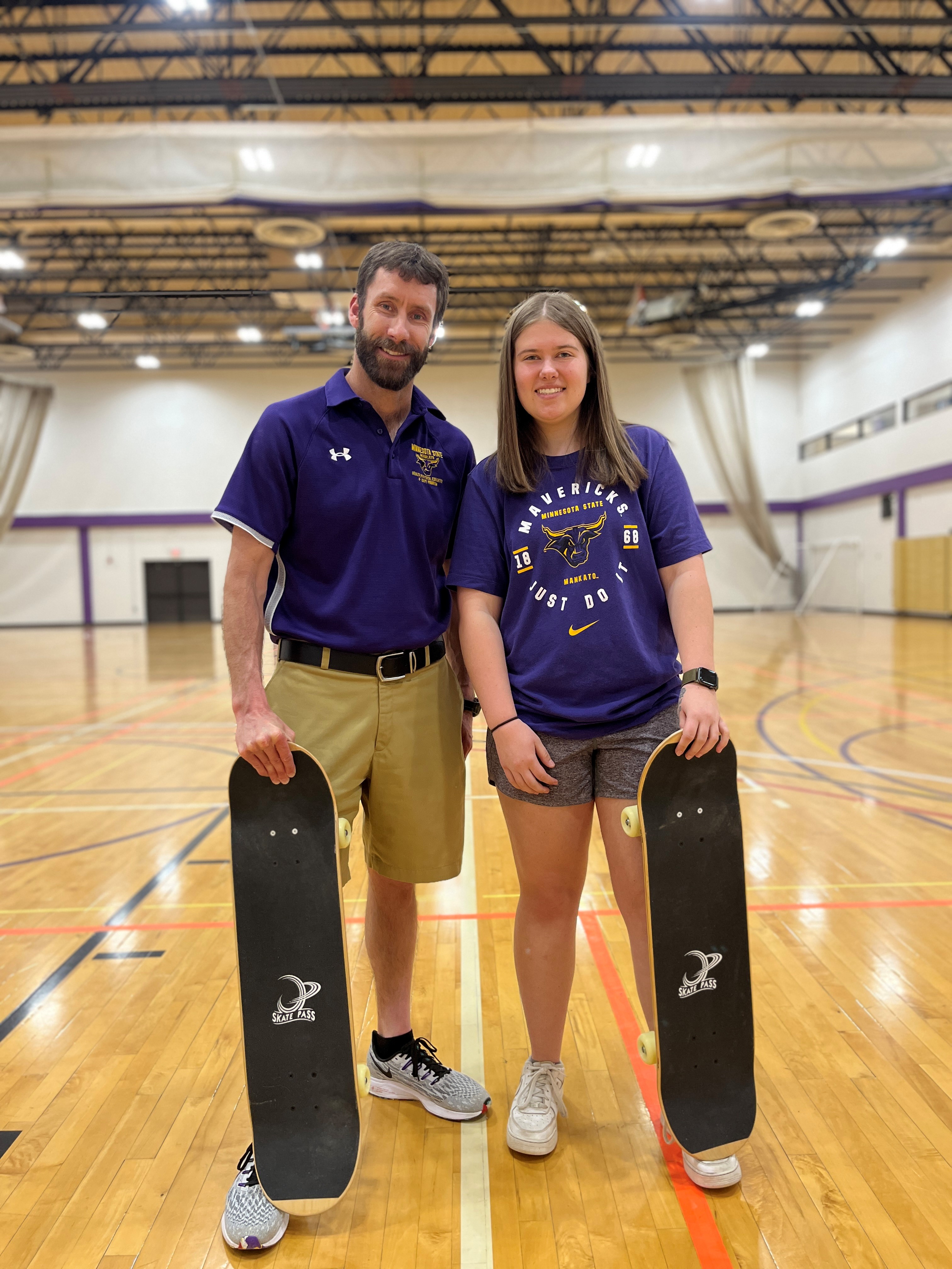 Professor Ben Schwamberger with student Mallory Stiff, in a gymnasium on campus, both holding skateboards.