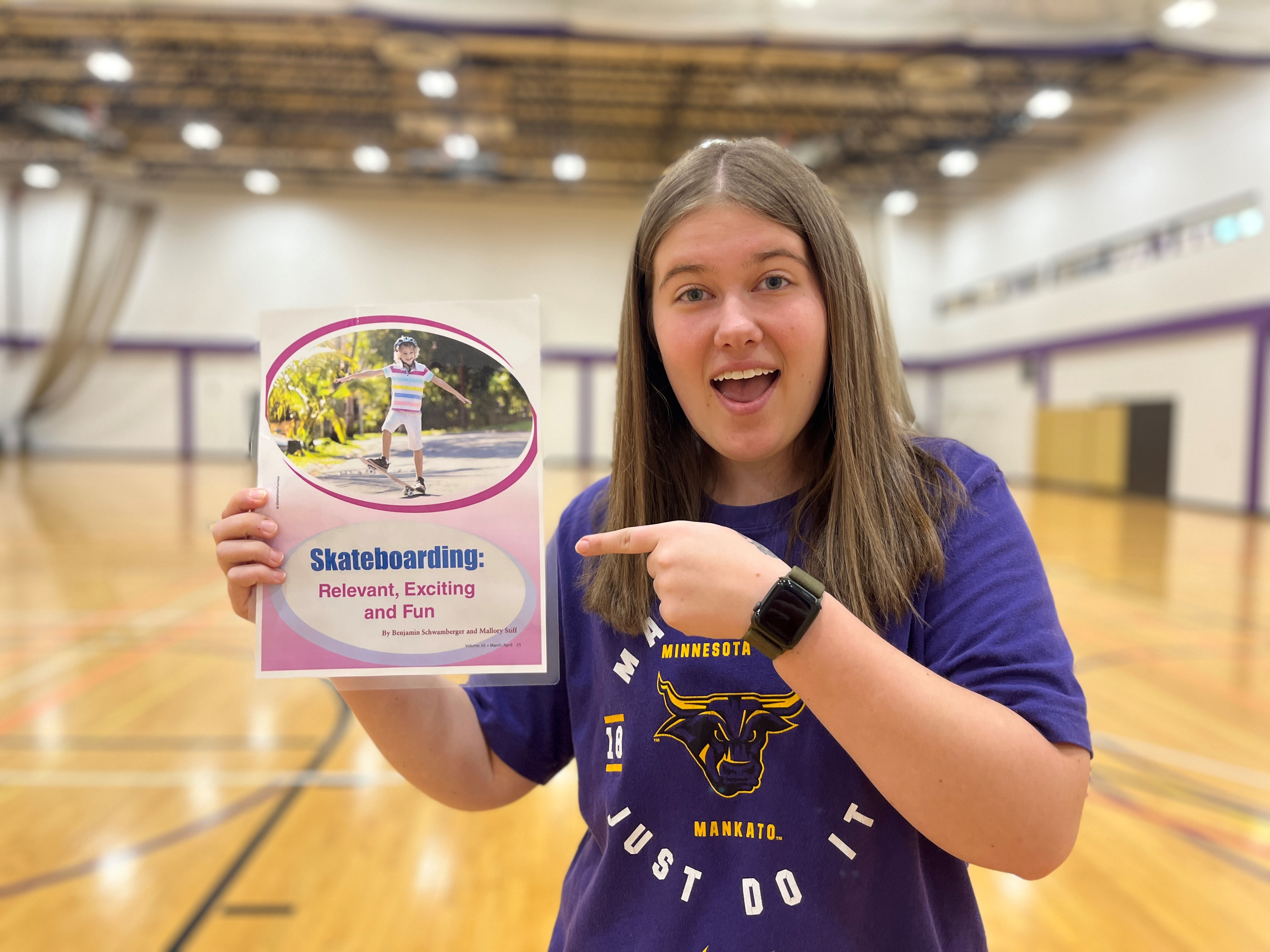 Mallory Stiff pointing to a journal she was published in. She is standing in a gymnasium.