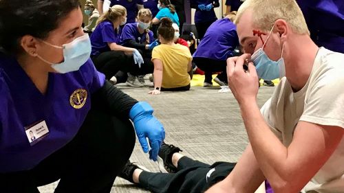 A nursing student in scrubs provides care to another student who is playing the role of someone with a head injury