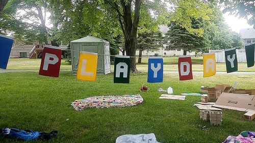 Banner hung between trees in a park that says Play Day with boxes and blankets on the ground