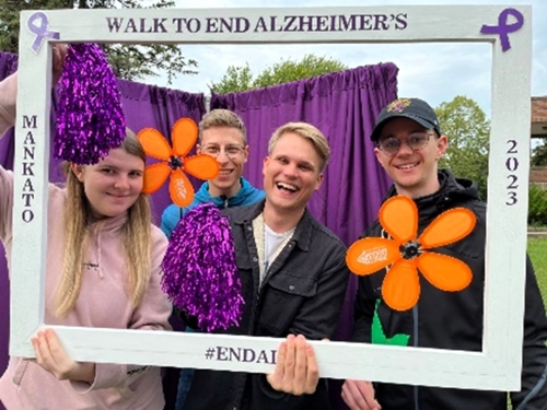 Four Austrian students posing outside holding up and looking through an empty picture frame with paper flowers at the Walk to End Alzheimer's event