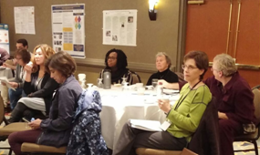 Attendees listening to a presentation at a table with research posters on the wall behind them 