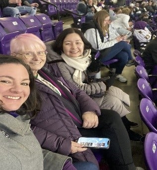 Three women smiling and sitting together in purple stadium seats at a sporting event