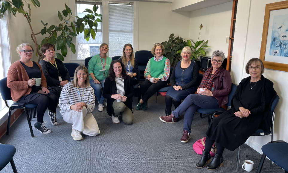 Group of ten women sitting in a circle in a casual meeting room, some holding mugs, smiling at the camera