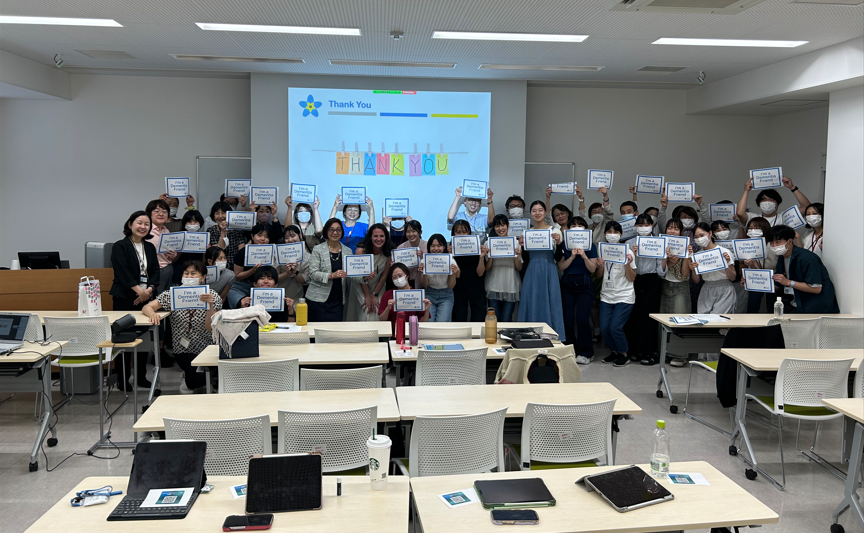 A group of people in a classroom, some kneeling in the front and others standing in the back, are holding up signs that say 