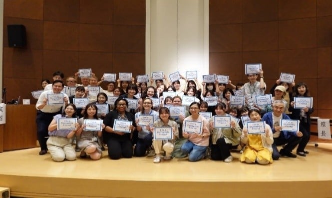 A large group of people on a stage is posing for a photo. They are holding signs with 