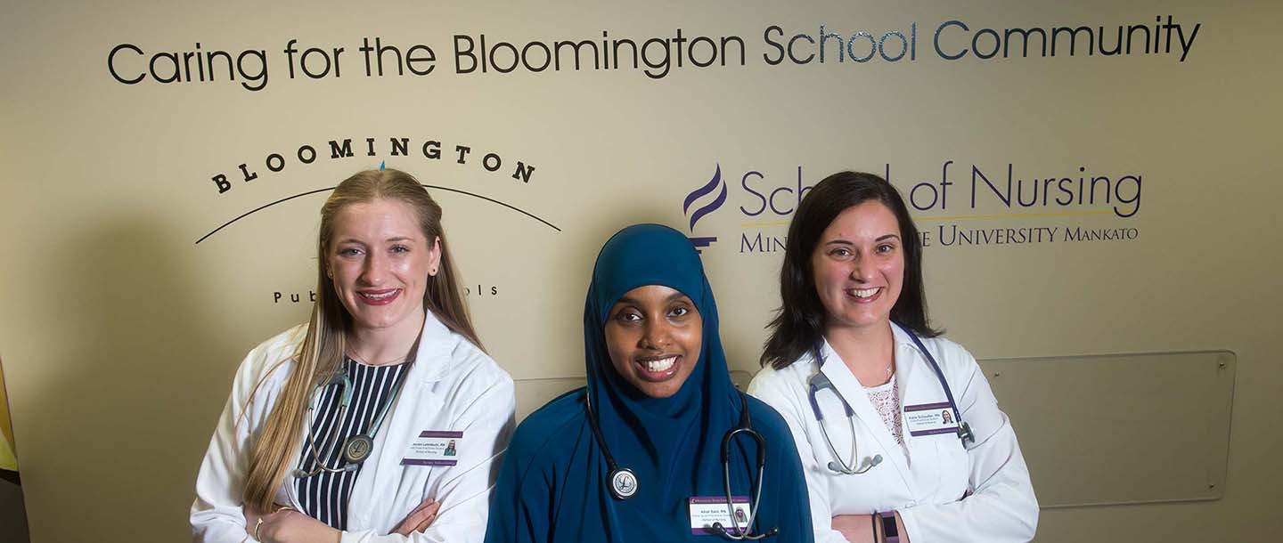 Three nurses posing in front of the Health Commons at Pond caring for the Bloomington school committee sign on the wall