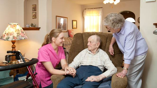 A nurse visiting an elderly couple in their home talking with the elderly man while holding his hand as he is sitting on a chair with his wife next to him