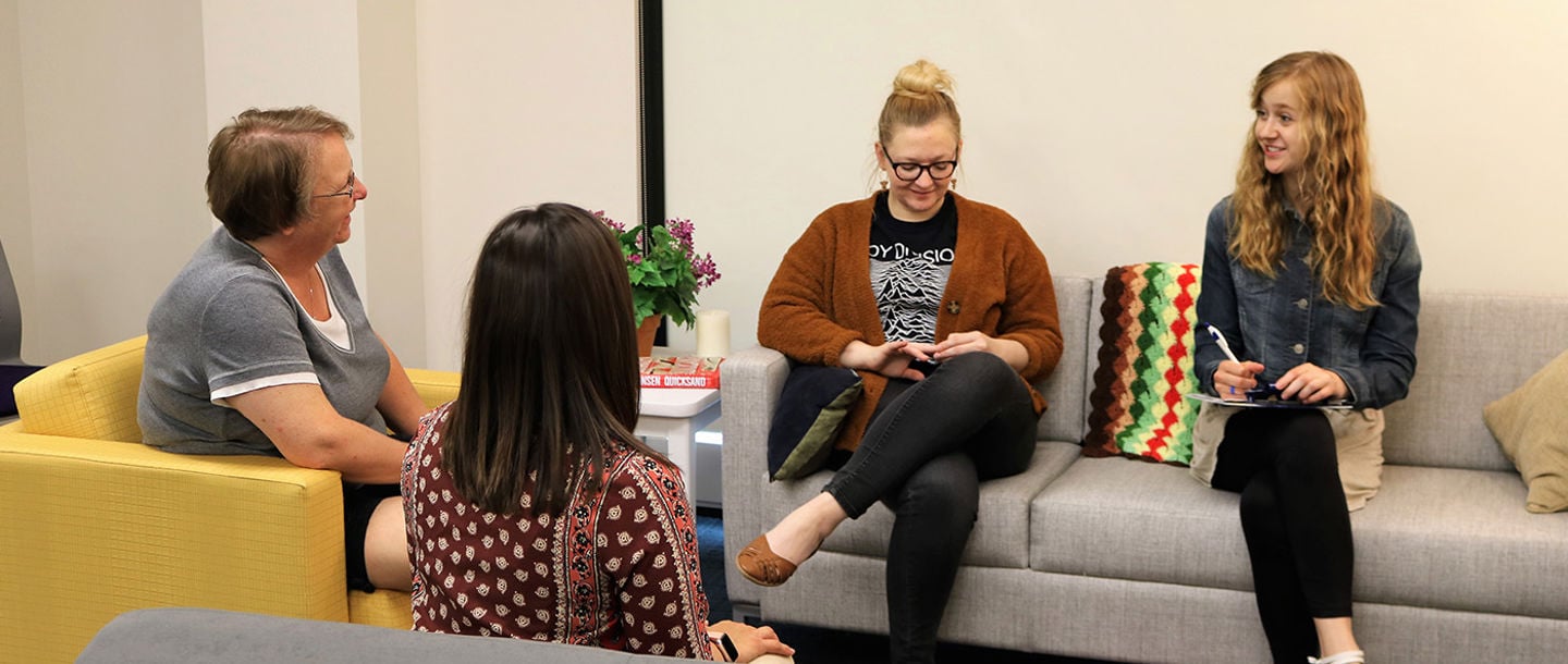 Three female students and professor of social work sitting on couches having a discussion while one student is taking notes