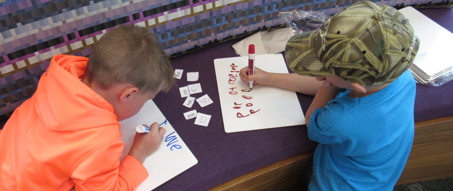 Two Rec N' Read kids one wearing a hoodie and another wearing a t-shirt with a cap writing on white paper using colored markers on a bench