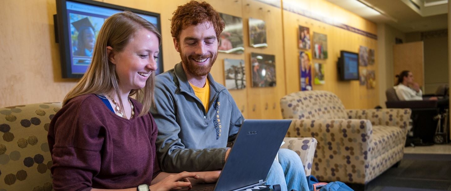 Two students, one male and one female working together on a laptop in a lounge area