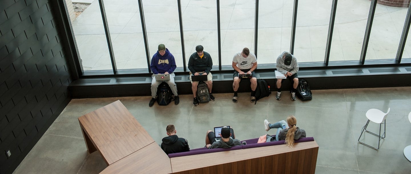 Seven students working on laptops, phones, and notes next to a big window